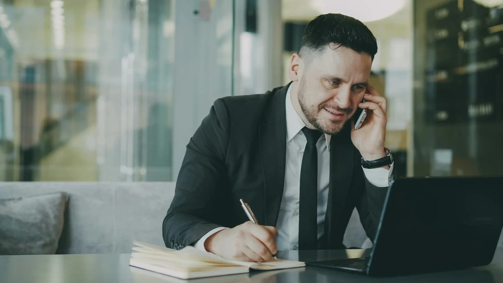 Telefonla görüşürken defterine not alan ve dizüstü bilgisayarla çalışan kişi